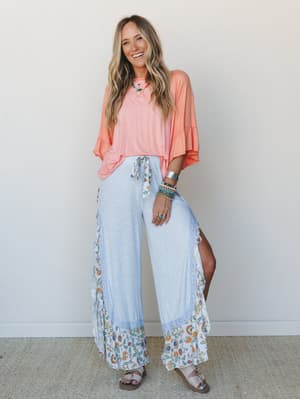 A smiling model in a breezy coral ruffle-sleeve top and wide-leg floral-trim pants styled with layered boho jewelry — the whole look reads like a really good Saturday with nowhere to be and everywhere to go.