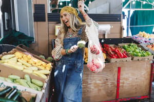 A laughing blonde woman shops a farmers market in denim overalls and a gingham shirt, holding up a mesh produce bag — the kind of outfit that feels equally at home picking tomatoes or meeting friends for coffee.