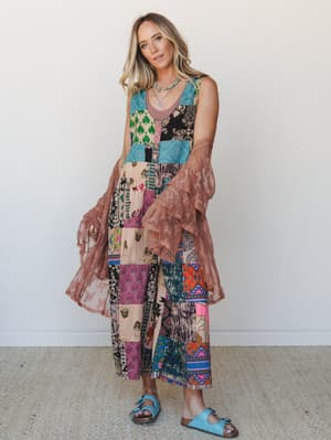 A patchwork maxi dress in a kaleidoscope of mixed prints is layered under a dusty rose lace duster with ruffle bell sleeves, grounded by blue buckle sandals — the kind of outfit that looks like it was collected over years and worn exactly right.