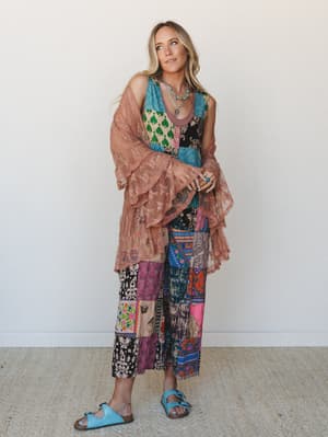 A free-spirited layered look built around a bold patchwork jumpsuit — worn with a dusty rose lace kimono draped over the arms and finished with turquoise sandals and layered silver jewelry for that effortlessly collected boho energy.
