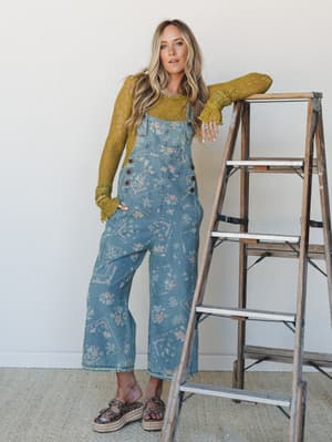 A sun-kissed blonde model leans casually against a weathered wooden ladder in floral-printed wide-leg denim overalls layered over a moss green lace long-sleeve top — the kind of outfit that feels equally at home at a farmers market or a backyard gath