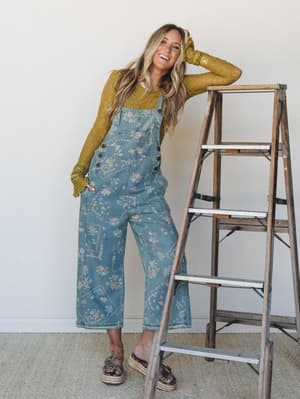 A laughing model leans against a weathered wooden ladder in floral-printed wide-leg denim overalls layered over a mossy green lace long-sleeve top — the kind of outfit that looks like it came together effortlessly but absolutely didn't.