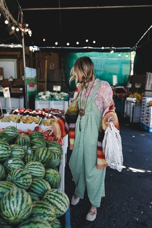 A model browses a farmers market in a green gingham wide-leg jumpsuit layered over a pink floral blouse, finished with a chunky striped crochet cardigan, woven flats, and a mesh tote — the whole look feels like a Sunday morning that got the lighting