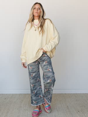An oversized butter-yellow sweatshirt layered over camo wide-leg jeans with a floral print overlay — the kind of outfit that feels effortless and a little unexpected, finished with rainbow platform sandals and layered necklaces.