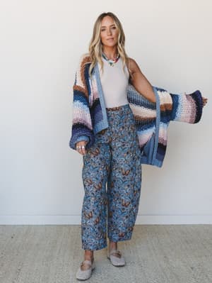 A relaxed full-body shot of a model wearing a butterfly-print wide-leg pant set paired with an oversized chunky stripe knit cardigan, layered with a colorful beaded necklace — the whole look has that effortless 'collected it slowly, wore it all at on