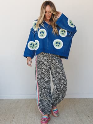 A bold, pattern-clashing outfit built around an oversized cobalt blue knit pullover with peace sign intarsia and wide-leg leopard print pants with a rainbow side stripe — the kind of look that has a whole personality before you even leave the house.