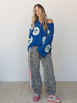 A relaxed, pattern-mixed outfit full of personality — an oversized blue peace sign knit sweater worn off-shoulder pairs with wide-leg leopard print jogger pants and rainbow platform sandals for a look that's equal parts retro and effortlessly fun.