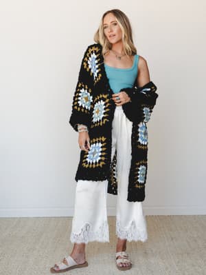 A longline granny square crochet cardigan in black with dusty blue, mustard, and white details layered over a teal tank and cream wide-leg satin pants with lace hem trim — the kind of outfit that looks effortless but clearly wasn't an accident.