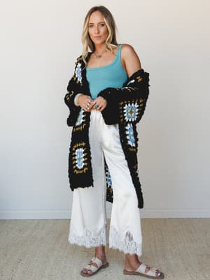A relaxed full-body shot of a model layering a chunky granny square crochet cardigan over a sky blue tank and wide-leg white satin pants with lace hem trim — the kind of effortless mix that feels intentional without trying too hard.