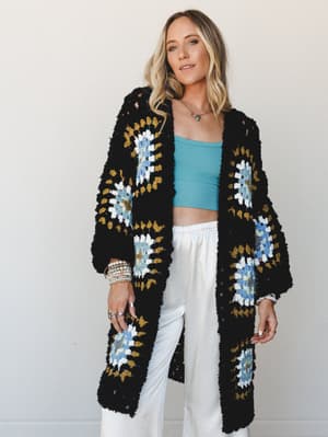 A long black granny square crochet cardigan styled over a sky blue crop top and white wide-leg pants — the kind of outfit that looks like it came together effortlessly but definitely did not.