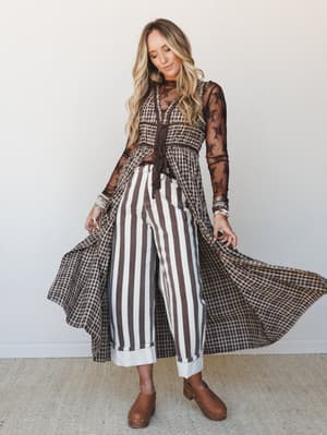 A layered three-piece look full of pattern-mixing energy — gingham maxi duster with sheer lace sleeves, striped wide-leg jeans, and clog boots, all in a rich chocolate and cream palette that feels effortlessly collected.