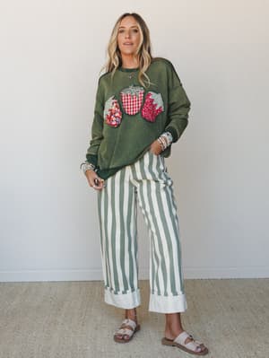 A relaxed, layered look built around an olive acid-wash sweatshirt with patchwork strawberry appliqués and wide-leg green-and-cream striped trousers with a cuffed hem — the kind of outfit that feels intentional without trying too hard.