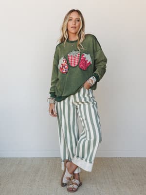 An olive mineral-washed sweatshirt with patchwork strawberry appliqués pairs with wide-leg cream and sage striped pants — the kind of outfit that looks effortless but is clearly doing a lot of good work.