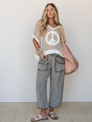 A relaxed, free-spirited look built around an open-knit crochet sweater with a peace sign graphic layered over wide-leg cargo pants in washed olive-grey, styled with a woven tote and sandals — the kind of outfit that looks effortless but definitely w