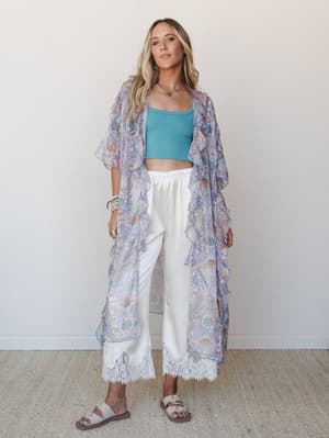 A flowy periwinkle floral kimono with ruffle trim layers over a cropped teal tank and white satin wide-leg pants finished with lace hem detail — the kind of outfit that feels equally at home at a summer brunch or wandering a seaside market.