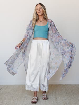 A flowy floral kimono in soft periwinkle and blush is layered over a teal crop top and wide-leg white pants with lace hem trim — the kind of outfit that feels effortless but clearly thought through.