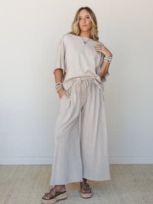 A matching mineral-washed taupe two-piece set — oversized raw-edge tee and wide-leg drawstring pants — styled with layered beaded bracelets, a turquoise pendant necklace, and platform espadrille sandals against a clean white wall.
