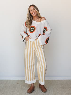 A smiling model in a playful, retro-inspired two-piece look — an oversized cream knit sweater with bold peace sign appliqués paired with wide-leg yellow striped pants and brown wooden clogs, shot against a clean studio wall with an easy, lived-in con