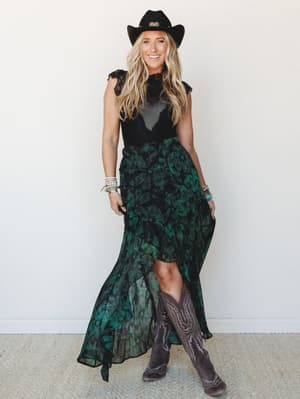 A smiling model in a moody black lace top and flowing black-and-green floral maxi skirt paired with tall cowboy boots and a black felt cowboy hat — the kind of outfit that shows up to a concert and stays through the after-party.