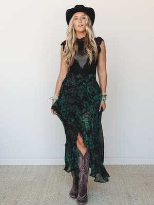 A flowing black and emerald floral maxi dress with lace detailing is styled with a wide-brim black hat and tall cowboy boots — the kind of outfit that walks into a room and makes it more interesting.