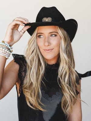 A close portrait of a model tipping a black felt cowboy hat with a gold concho detail, styled with a black lace top and stacked beaded bracelets — the whole look has that effortless western-boho energy that feels like a concert and a campfire at once