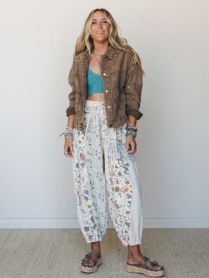 A smiling blonde model layers a washed mocha denim jacket with star embellishments over a teal crop top and voluminous floral patchwork pants, styled with platform sandals and stacked beaded bracelets — the whole look has that effortless, 'I threw th