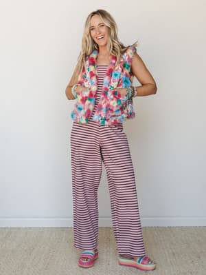 A full-body studio shot of a smiling model in a pink-and-black striped wide-leg jumpsuit layered under a multicolor floral faux fur vest, finished with rainbow platform sandals and stacked bracelets — the whole look reads like a color run and a good