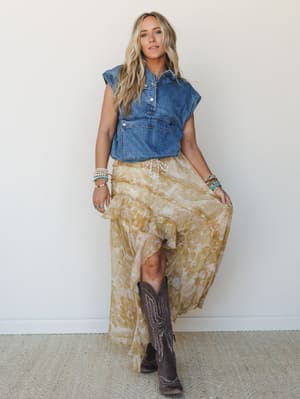 A breezy full-body shot of a model pairing a cropped sleeveless denim top with a flowy floral maxi skirt in golden yellow, finished with brown cowboy boots and stacked turquoise and beaded bracelets — the kind of outfit that looks like it was thrown