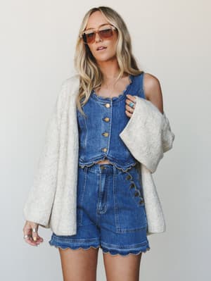 A denim two-piece set — scallop-trimmed crop top and matching high-waist shorts — layered under an oversized cream knit cardigan, styled with retro tinted sunglasses and stacked silver and turquoise rings for an easy, sun-warmed boho feel.