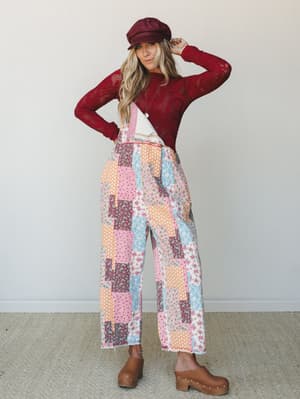 A full-body model shot of a layered boho look — patchwork wide-leg overalls in mixed floral prints styled over a deep burgundy crochet long-sleeve top, finished with a newsboy cap and wood-studded clogs for a vintage-market kind of charm.