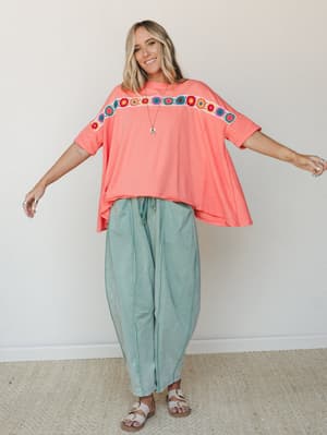 A smiling model in a breezy coral oversized tee with a bold crochet floral trim across the chest, paired with relaxed sage green drawstring pants and white sandals — the kind of outfit that feels like a good mood.