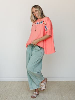 A coral oversized tee with colorful crochet floral trim across the chest is paired with sage green wide-leg drawstring pants and white buckle sandals — casual, colorful, and the kind of outfit that feels just as good as it looks.
