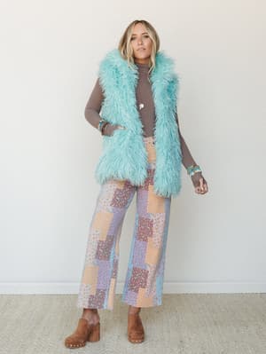 A long mint faux fur vest layered over a mocha turtleneck and wide-leg patchwork pants in pastel florals — the kind of outfit that makes people stop and ask where you got those pants.