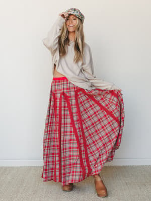 A smiling model in a flowing red plaid maxi skirt, cozy oatmeal sweatshirt, and tan ankle boots — the kind of outfit that looks like it was thrown together perfectly on a crisp fall morning.
