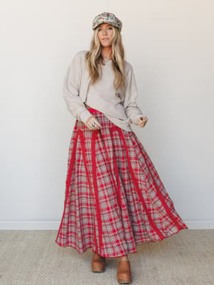 A relaxed full-body model shot of a red plaid maxi skirt styled with an oatmeal crewneck sweatshirt, floral baker boy cap, and tan wooden clogs — the kind of outfit that feels vintage-found but totally intentional.