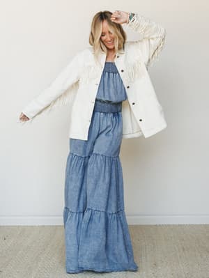 A smiling model in a chambray tiered maxi dress layered under a cream fringe jacket, shot against a soft neutral wall — the kind of outfit that feels equally at home at a summer concert or a breezy weekend brunch.