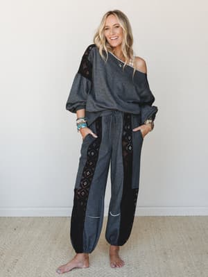 A smiling model wears a matching charcoal two-piece set with black lace panel inserts — the oversized off-shoulder top and relaxed jogger pants styled with stacked turquoise bracelets and layered necklaces, shot against a clean neutral interior.