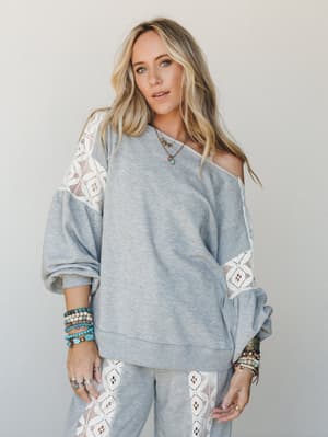 A relaxed heather grey sweatshirt-and-jogger set with geometric crochet lace paneling along the sleeves and pant sides, styled off-the-shoulder and stacked with turquoise and mixed-metal jewelry for that effortless boho-at-home feel.