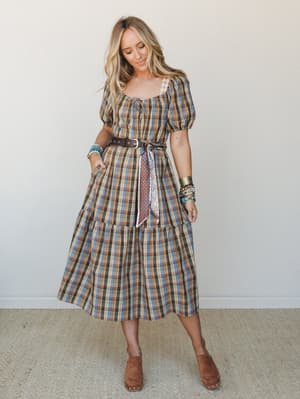 A plaid midi dress with puff sleeves and a tiered skirt, styled with a leather belt, knotted scarf, and stacked bracelets — the kind of outfit that looks intentional without trying too hard.