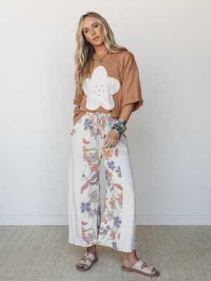 A breezy two-piece set — a mocha oversized tee with a white flower appliqué patch and wide-leg floral pants in cream — styled with layered turquoise bracelets and buckle sandals for that effortless, lived-in boho feel.