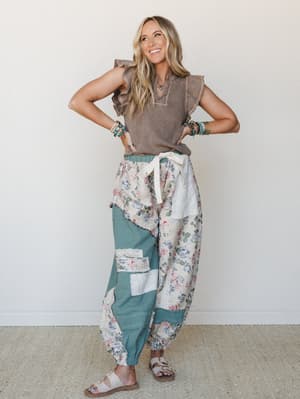 A smiling model pairs a mocha ruffle-sleeve top with voluminous patchwork floral jogger pants in sage and cream, styled with layered boho jewelry and flat sandals — the whole look reads like a really good thrift find you actually planned.
