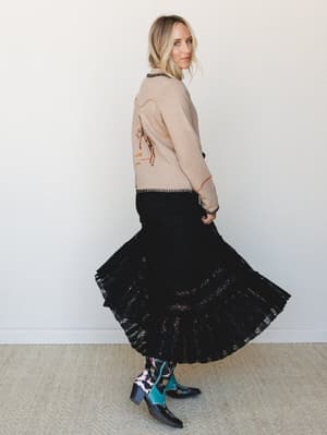 A model shot in profile against a clean white studio wall, wearing an embroidered western sweater layered over a flowing black lace maxi skirt, finished with colorful embroidered cowboy boots — the kind of outfit that makes you want to twirl on purpo