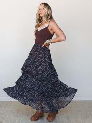 A tiered polka dot maxi skirt swirls mid-motion against a clean studio backdrop, paired with a burgundy lace-trim camisole and wood-heel clogs — the kind of outfit that looks just as good twirling at a farmers market as it does at a fall dinner with