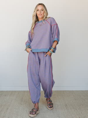 A matching striped sweatshirt and jogger set in dusty blue with coral-red pinstripes, styled with stacked turquoise bracelets and espadrille platform sandals against a clean neutral wall — relaxed and put-together at the same time.