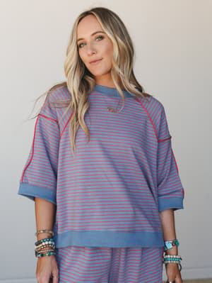 A relaxed striped sweatshirt top shot mid-body against a soft neutral wall — the kind of effortless two-piece set that feels like a good mood in fabric form, styled with layered boho bracelets and a delicate 'mama' necklace.