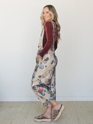 A smiling model poses in profile wearing a floral wide-leg overall jumper layered over a rich burgundy velvet long-sleeve top, styled with platform espadrille sandals and a turquoise beaded bracelet — the kind of effortless fall outfit that looks lik