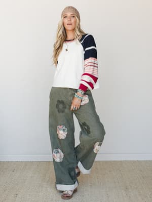 A relaxed, personality-forward outfit pairing a color-blocked striped sweater with wide-leg olive jeans covered in floral appliqués and tonal daisy prints — the kind of look that feels effortlessly collected, like she just grabbed what made her happy