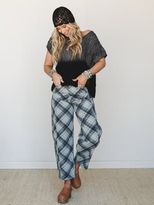 A smiling model in a relaxed full-body shot layers a cable-knit dip-dye sweater top with wide-leg plaid pants, a black crochet beanie, and tan clogs — the kind of effortless mix-and-match energy that makes the outfit feel personal rather than put-tog