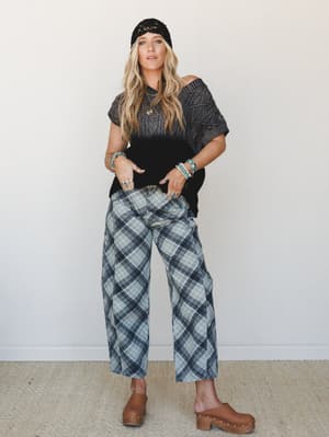 A relaxed, layered look with a cable-knit oversized sweater top styled off-shoulder over wide-leg plaid pants in sage and navy, grounded by tan clogs and accessorized with stacked turquoise jewelry and a black crochet beanie — the kind of outfit that