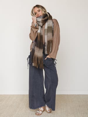 A relaxed, layered fall outfit — oversized camel top, wide-leg dark wash pants with drawstring side details, and a chunky fringed plaid scarf wrapped up to the chin, styled with flat sandals and stacked jewelry for that effortlessly cozy, put-togethe